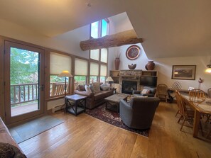 A spacious living area with a fireplace, wooden furniture, a dining table with chairs, and large windows offering a view of the outdoors.