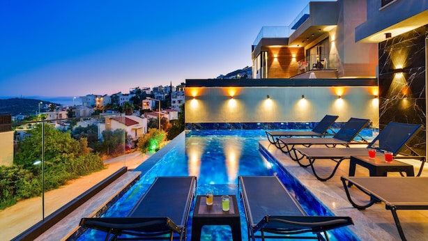 A rooftop infinity pool with lounge chairs and a table, overlooking a cityscape at dusk. - Sun, sea, and serenity — your perfect Kalkan getaway (Kalkan Centre)