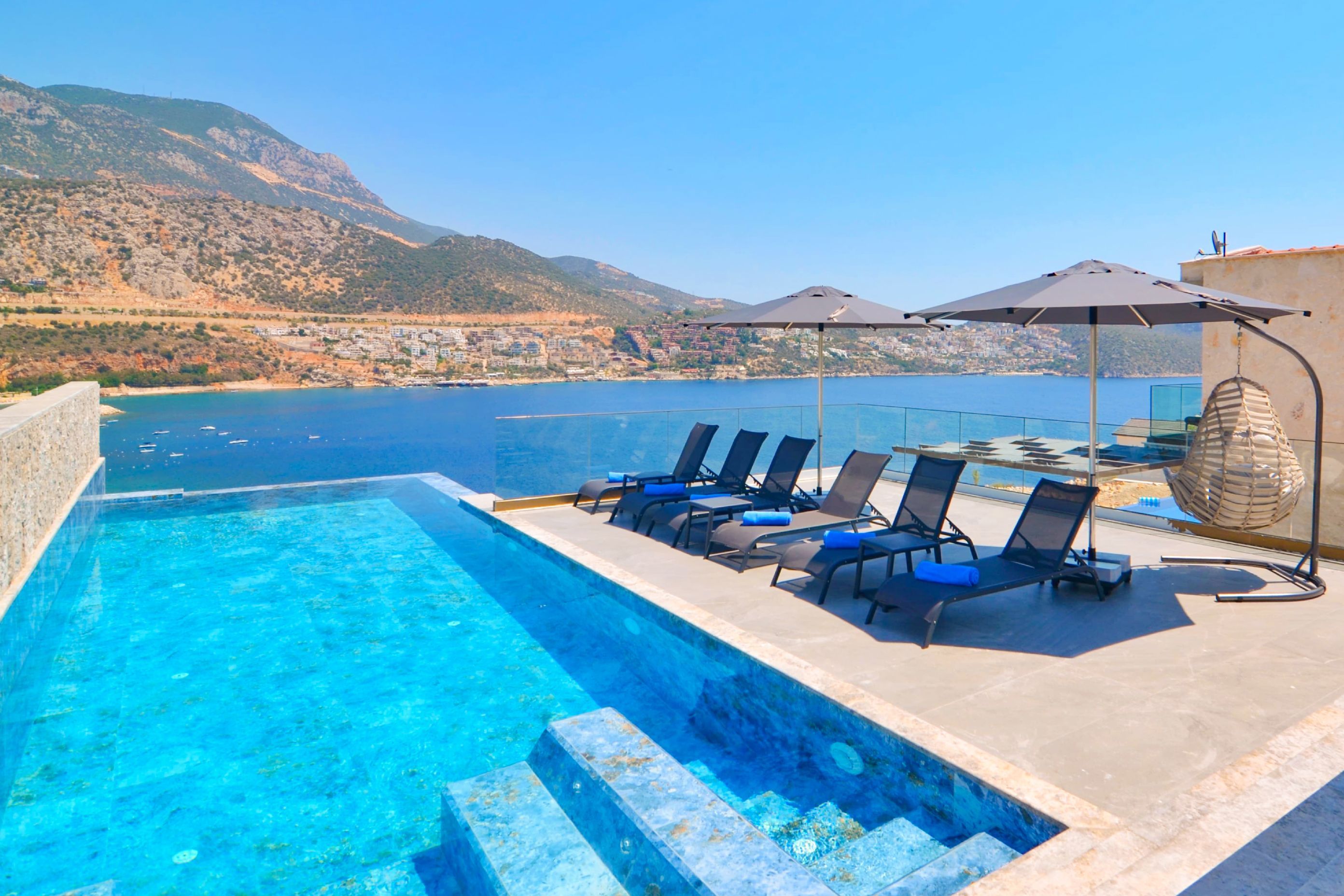 Une magnifique piscine à débordement d’un bleu profond, accessible par des marches, entourée de chaises longues et de parasols, et offrant une vue imprenable sur une ville côtière et les montagnes.