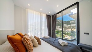 A modern bedroom with a large bed, multiple pillows, a bench, and a view of a mountainous landscape through a glass door. - Stylish Villa with Private Pool & Sea Views (Kalkan)