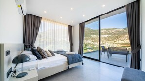 A modern bedroom with a large bed, a bedside table with a lamp, a view of a balcony with outdoor furniture, and a cityscape in the background. - Stylish Villa with Private Pool & Sea Views (Kalkan)