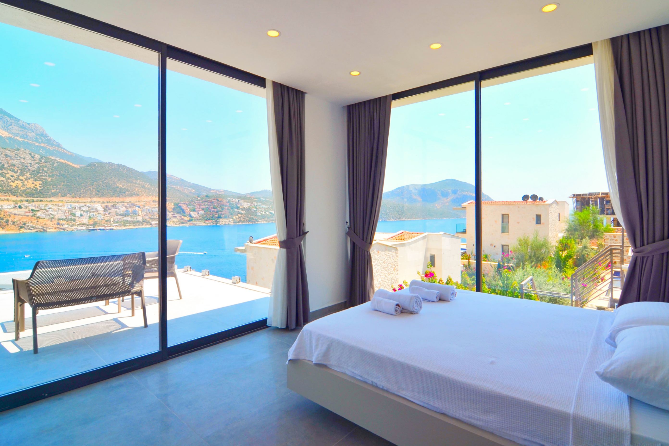 A hotel room with a large bed, a balcony with a table and chairs, and a view of the sea and mountains.