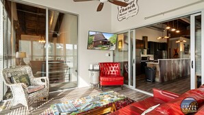 Un salón amplio con un sofá rojo, una silla de mimbre, una mesa de centro con revistas, un ventilador de techo y una cocina visible a través de puertas de cristal.