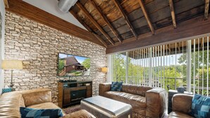 Una sala de estar acogedora con paredes de piedra, sofás de cuero, un televisor de pantalla plana y un ventanal con vistas al verdor.