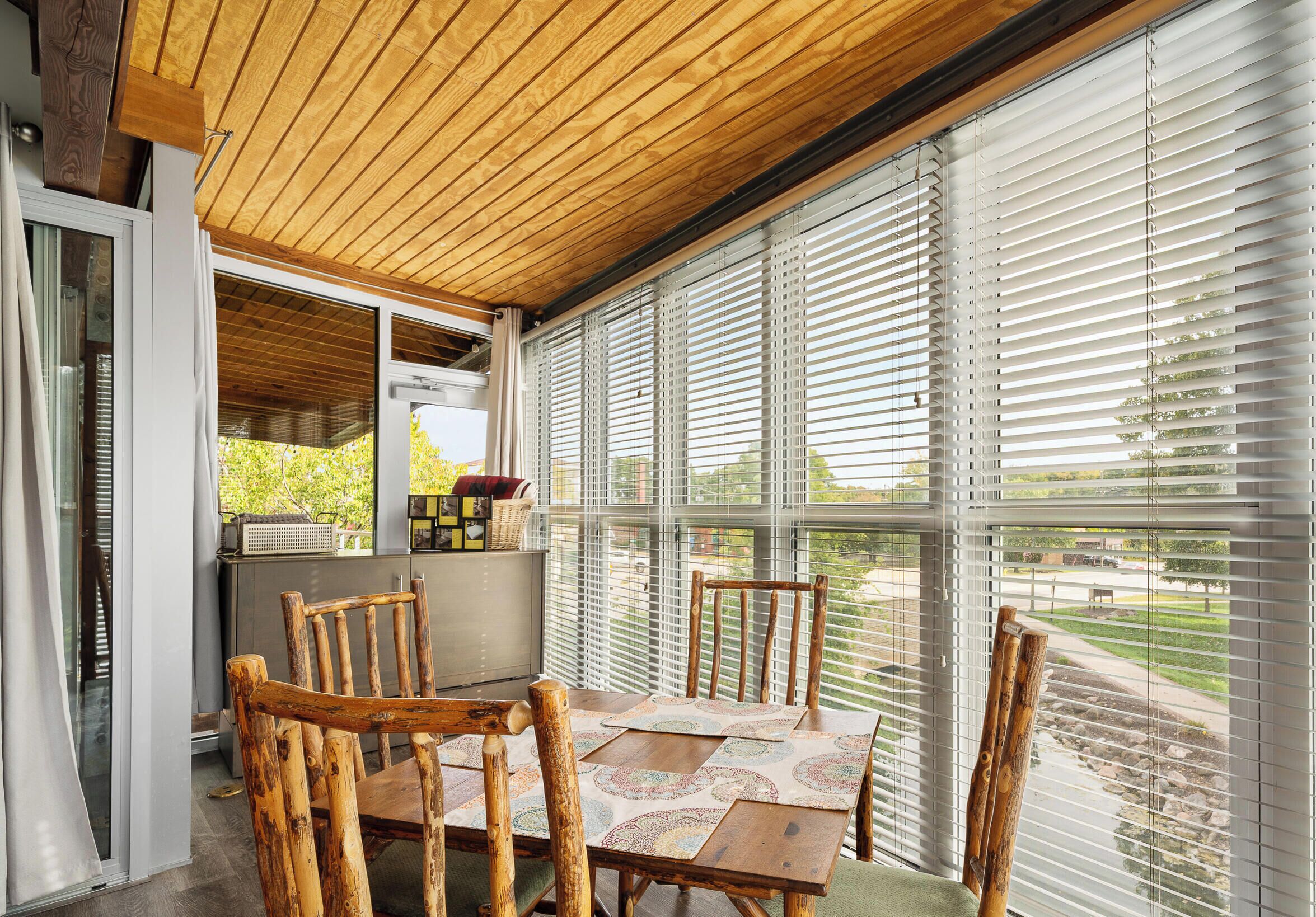 A dining area with a wooden table and chairs, large windows with blinds, and a view of the outdoors.