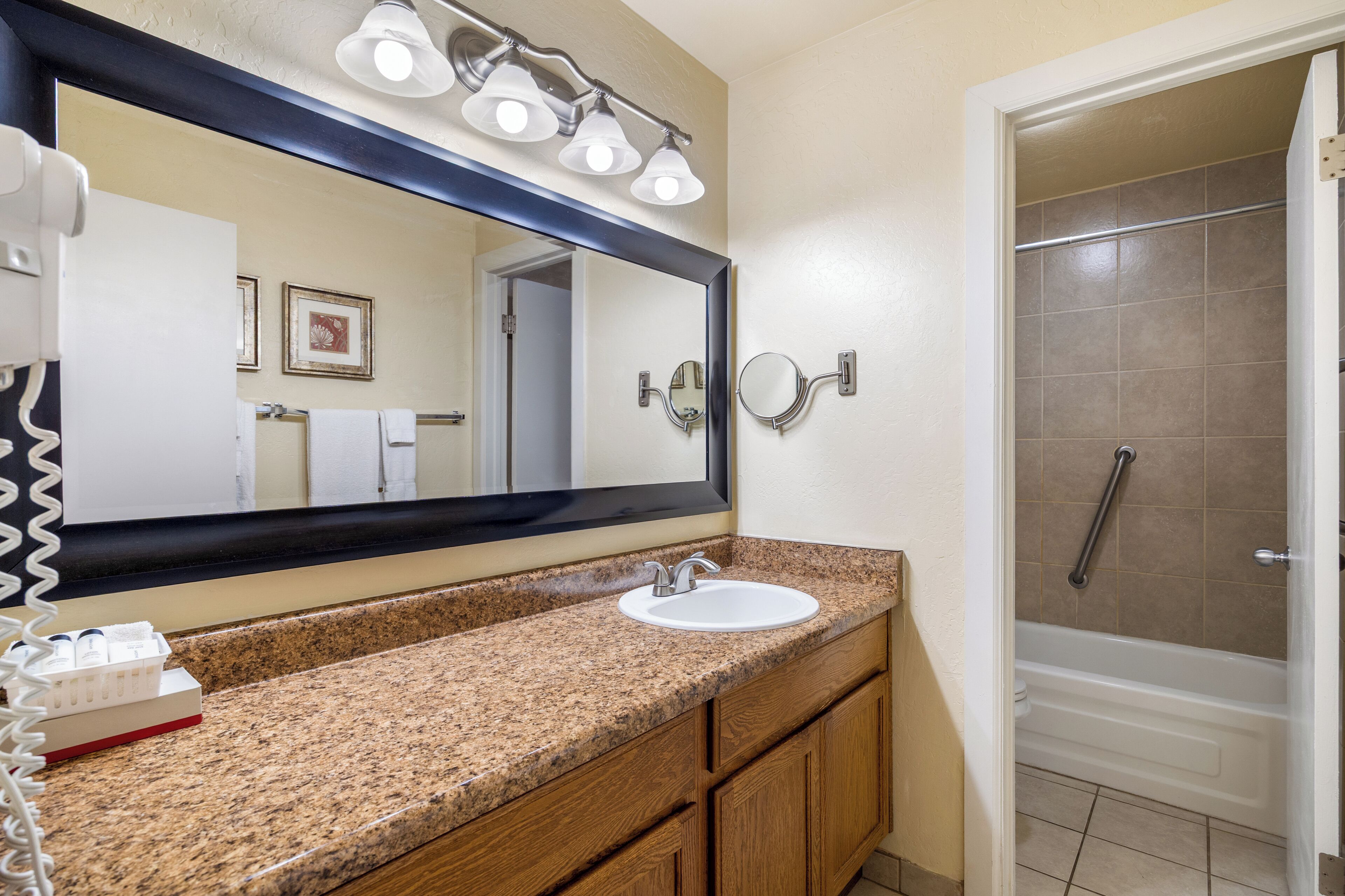 A bathroom with a granite countertop, white sink, and a large mirror. There is a shower area visible to the right.