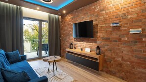 A modern living room with a brick wall, a flat-screen TV mounted above a wooden shelf, a blue sofa, and a small round table. - Spodek Modern Apartment (Katowice)
