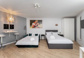A modern hotel room with two beds, a desk, a chair, a small table, a wall-mounted TV, and a ceiling light fixture. - Kraków Grzegórzki Deluxe Studio (Kraków)