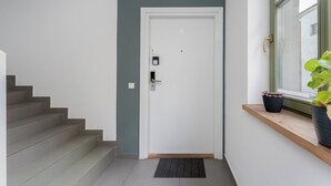 A hallway with a white door, a metal intercom, and a window with a plant. - Kraków Grzegórzki Deluxe Studio (Kraków)
