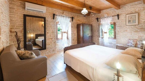 Um quarto com uma cama grande, teto de madeira, paredes de pedra, um espelho e uma janela com cortinas.