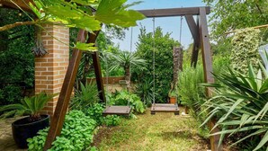 Um jardim com baloiço de madeira, parede de tijolos e diversos vasos com plantas.