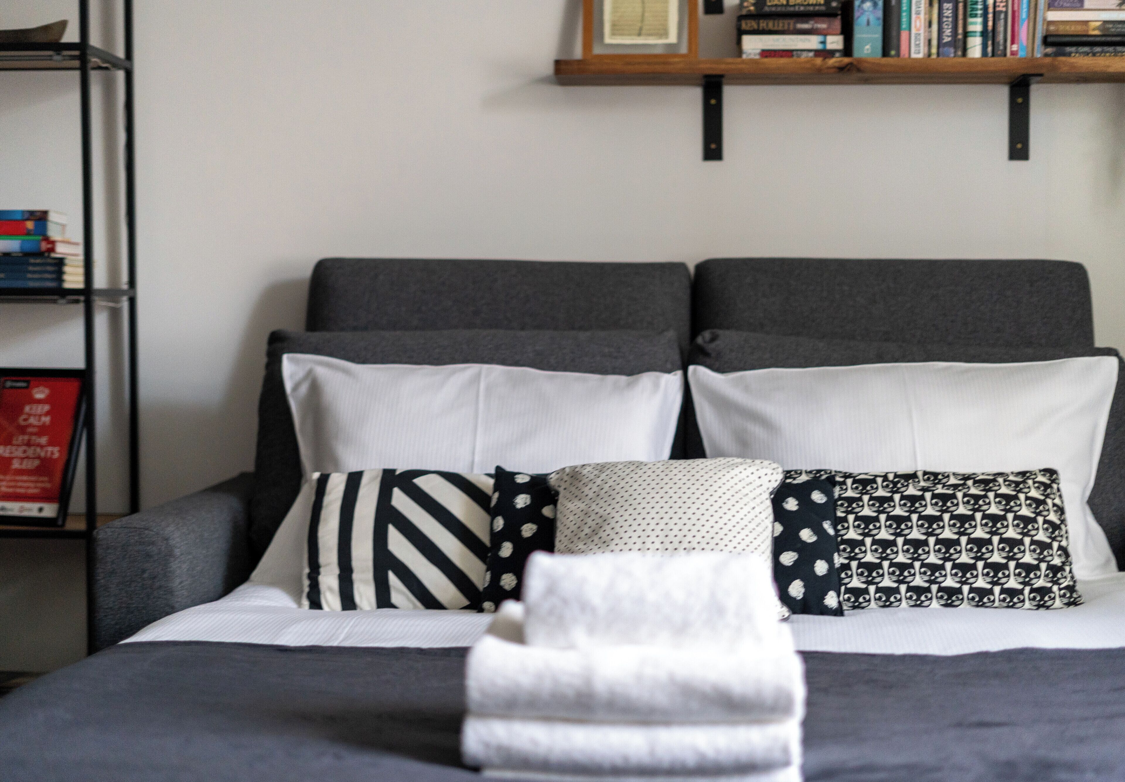 Una cama ordenada con cabecera gris, almohadas blancas y estampadas, y una toalla blanca doblada. Se ve una estantería con libros y un cuadro enmarcado al fondo.