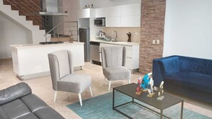 Een moderne woonkamer met een bank, twee fauteuils, een salontafel en een blauwe bank. In de achtergrond is een keuken met witte kasten en een magnetron te zien.