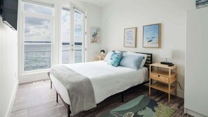 A bedroom with a bed, two bedside tables, a window with a view of the sea, and two framed artworks on the wall. - Chic Lakefront Getaway W/ Hot tub + Kayaks! (Alanson)