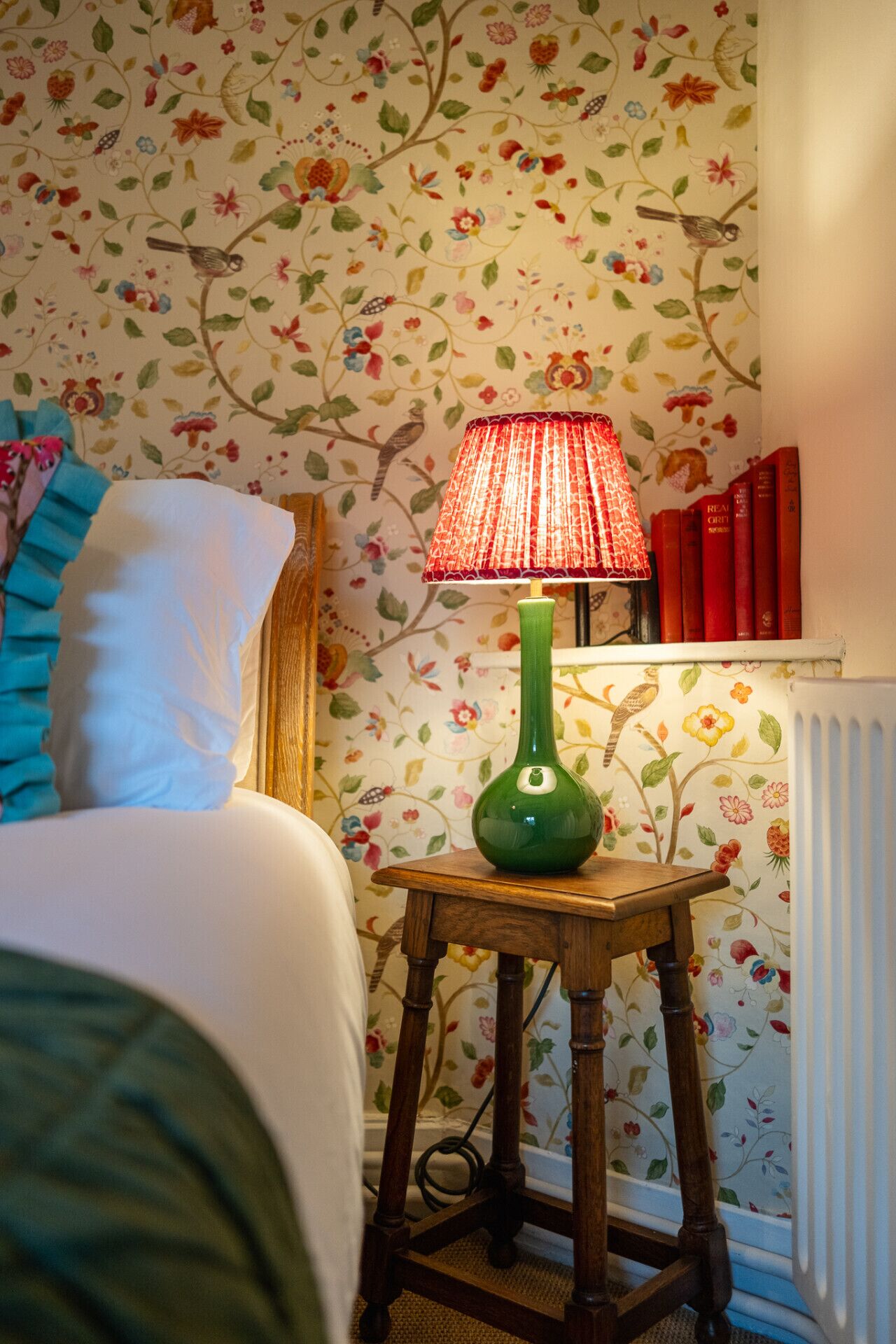 Een slaapkamer met bloemenbehang, een groene vazenlamp en een nachtkastje met boeken.