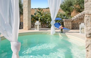 A pool area with a clear blue pool, white drapes, stone pillars, and a blue umbrella. - Lovely home in Modica (Modica)