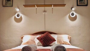 A neatly made bed with rolled towels, a wooden shelf above, and two wall-mounted lights. - Hostal Tres Reyes (Cusco)