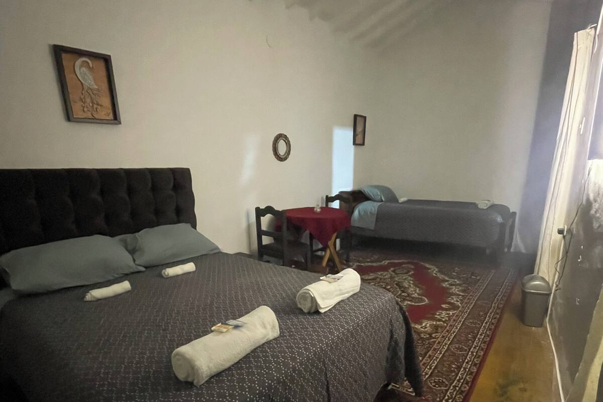 Een slaapkamer met een groot bed, een kleiner bed, een rood vloerkleed en een plafondventilator.