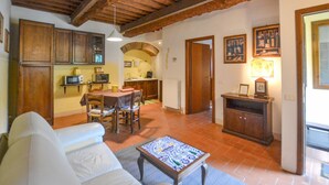 Uma sala de estar acolhedora com sofá, mesa de centro e área de jantar com cadeiras e mesa. Há uma cozinha com armários e micro-ondas. O quarto possui vigas de madeira no teto e piso de azulejos.
