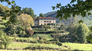 Um edifício de pedra com várias janelas e um telhado vermelho, rodeado por vegetação exuberante e colinas.