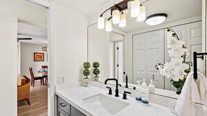 Un baño moderno con un lavabo de mármol blanco, grifo negro y un espejo grande con tres luces. Hay dos plantas en macetas y una orquídea blanca en la encimera.