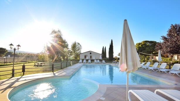 Ein Swimmingpool mit sonnendurchflutetem Wasser, umgeben von Liegestühlen und Sonnenschirmen, mit einem Gebäude und Bäumen im Hintergrund.