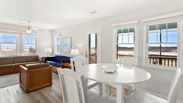 A spacious living room with a blue sofa, wooden furniture, a round dining table, and large windows offering a beach view.