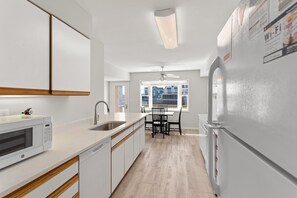 A modern kitchen with a microwave, sink, and refrigerator. There is a dining area with a table and chairs, and a window providing a view of the outside. - Egret – Bright Buxton Townhome w/ Ocean Views (Buxton)