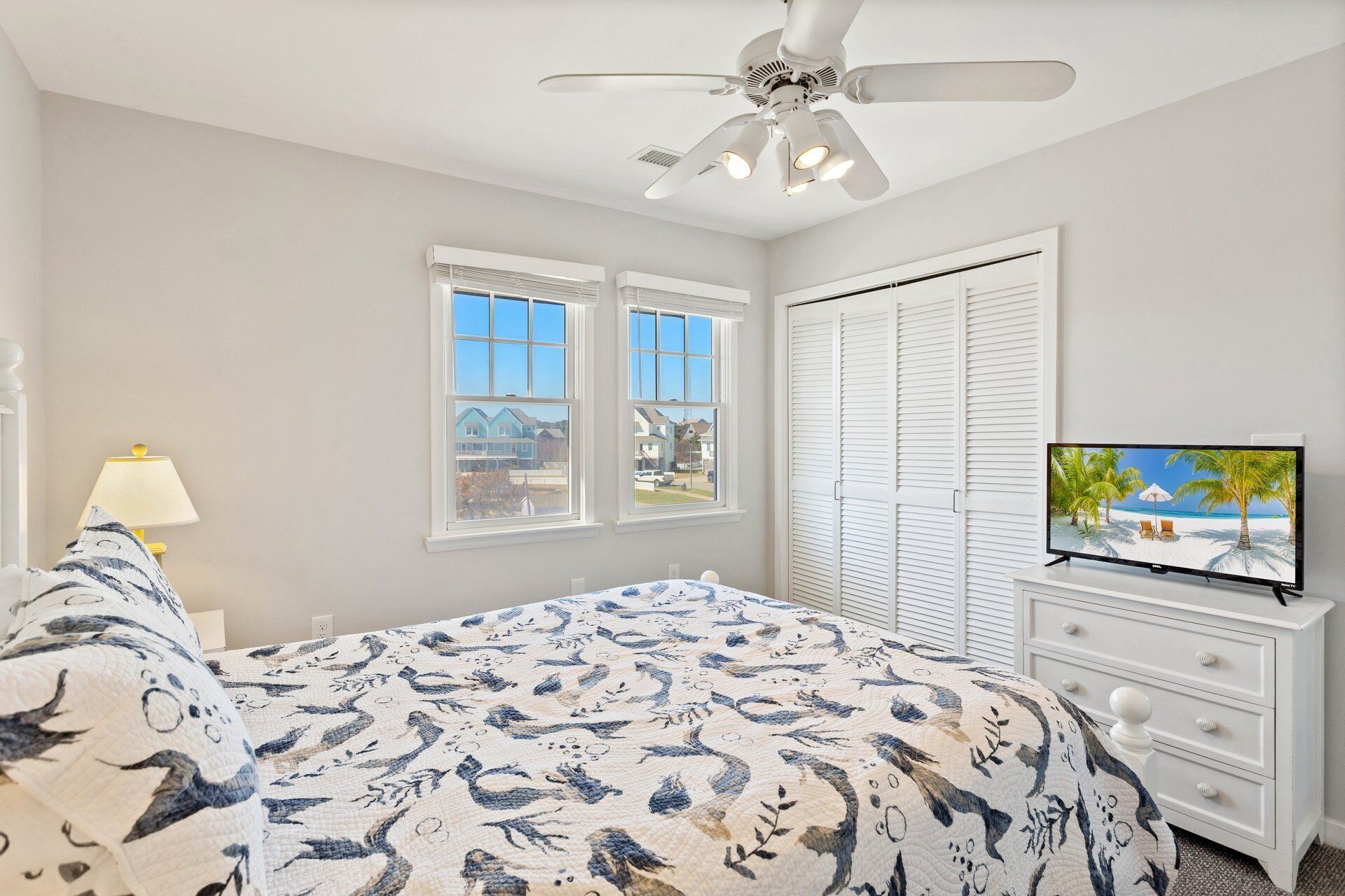 A bedroom with a bed, a TV, a ceiling fan, and two windows.