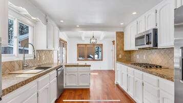 A modern kitchen with white cabinetry, granite countertops, a central island, and a view of the outdoors through a large window.