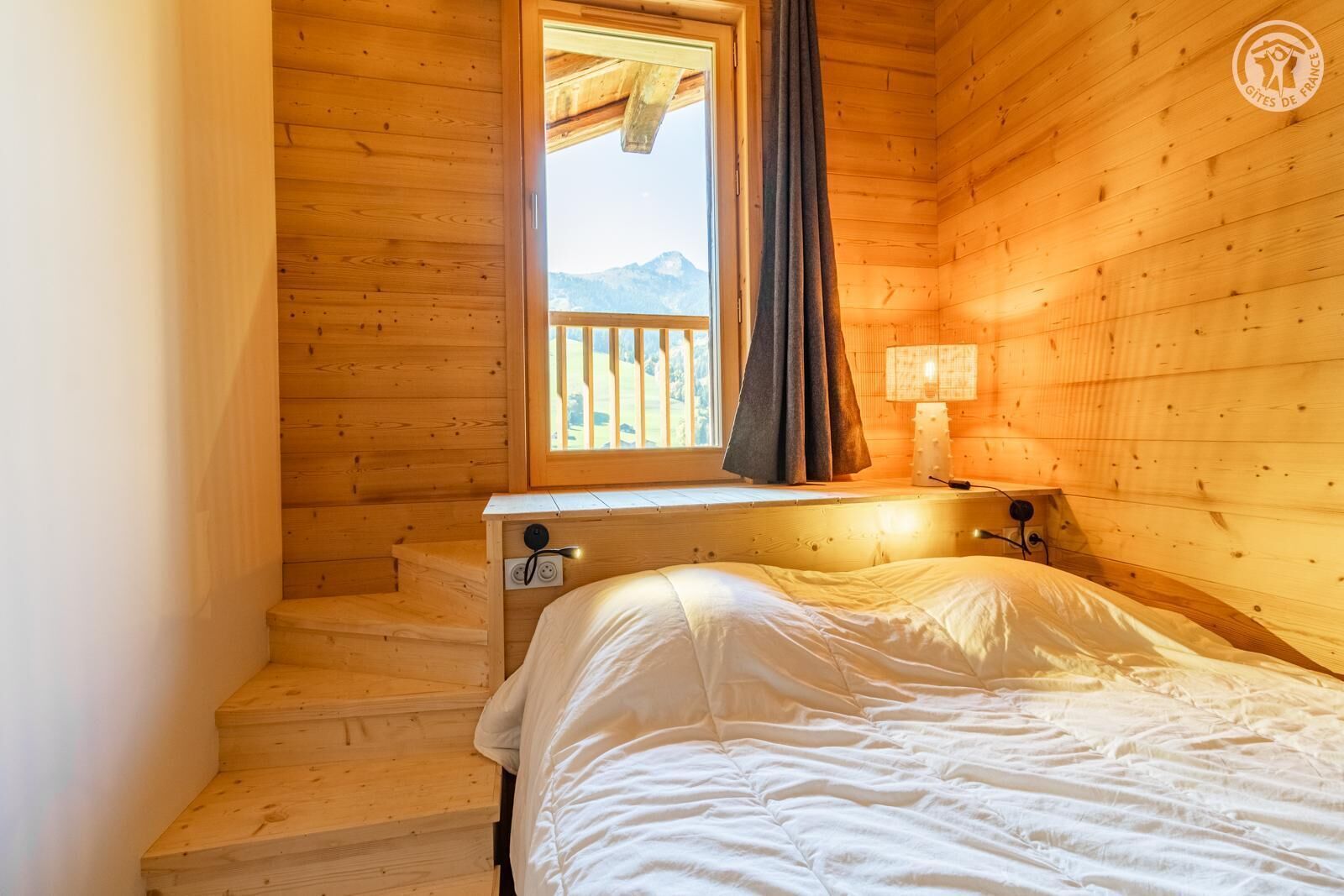 Ein Zimmer in einer Holzhütte mit einem Bett, einem Fenster mit Vorhängen, einem Nachttisch und einer Lampe.
