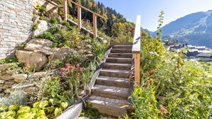 Un escalier en pierre, agrémenté de rampes en bois, mène au sommet d’une colline, entourée d’une végétation luxuriante, et offre une vue sur une vallée parsemée de bâtiments.