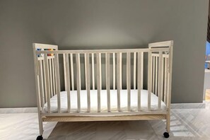 A white crib with a wooden frame, positioned against a grey wall. - EATON BY FEEL HOME (Kuala Lumpur)
