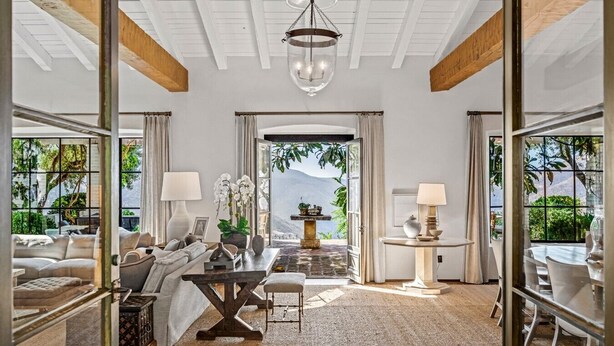 Une pièce spacieuse avec un plafond haut, un plafond en bois, un lustre, un coin repas avec une table et des chaises, un ensemble de canapés et une vue sur la verdure grâce à de grandes fenêtres.