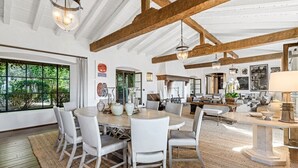 Un vaste espace de vie doté d’un plafond avec une grande poutre en bois, d’une table à manger avec des chaises, et offrant une vue sur l’extérieur grâce à de grandes fenêtres.