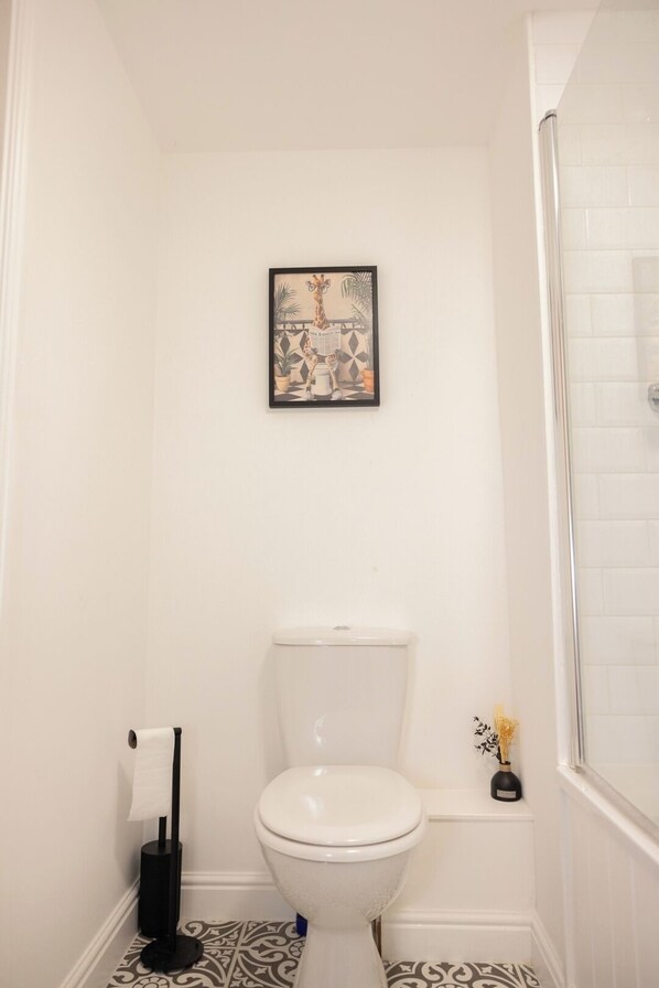 A bathroom with a toilet, a framed picture on the wall, a toilet paper holder, and a small vase with flowers.