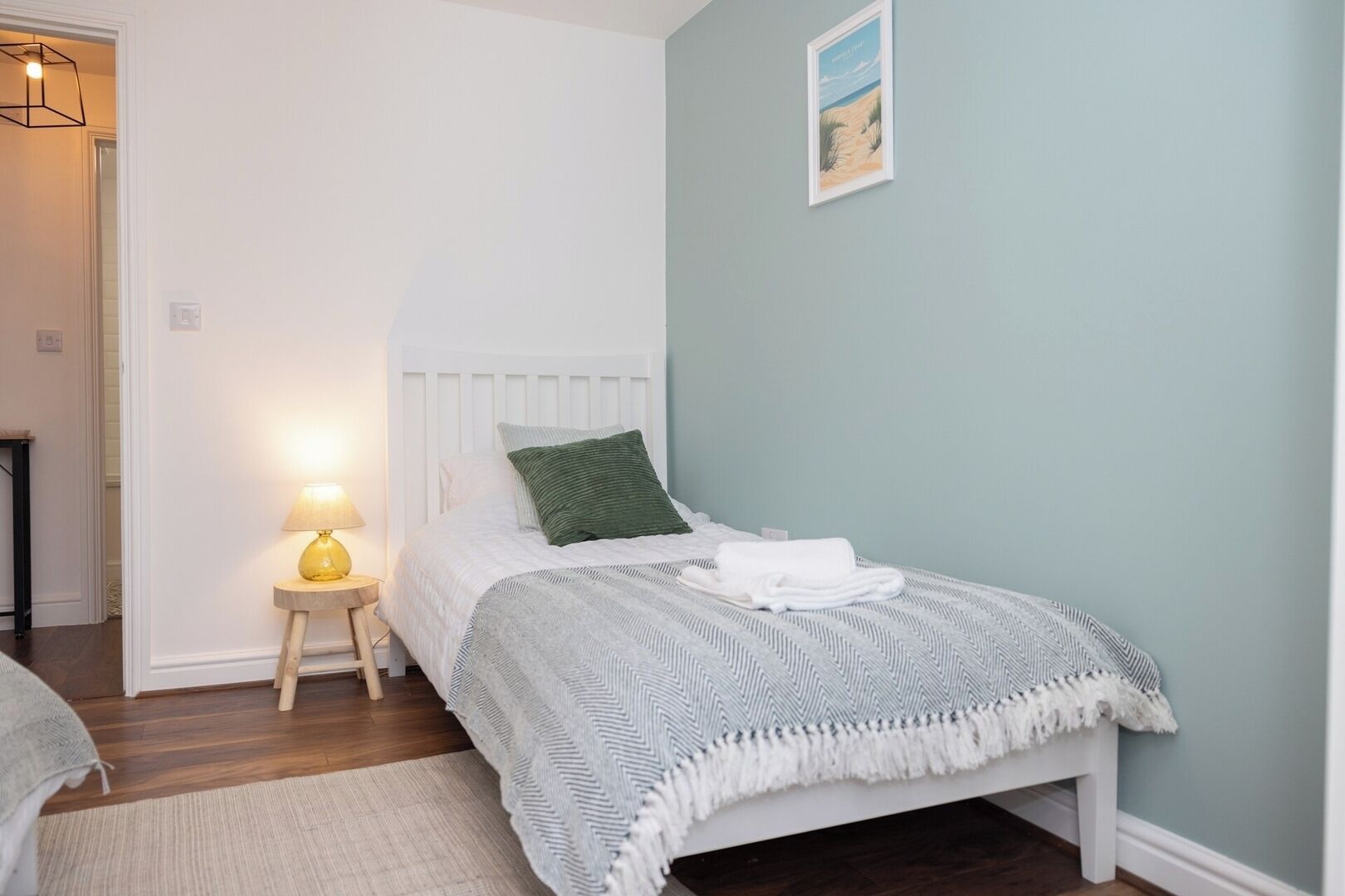 A bedroom with a bed, a bedside lamp, a framed picture on the wall, and a door leading to another room.