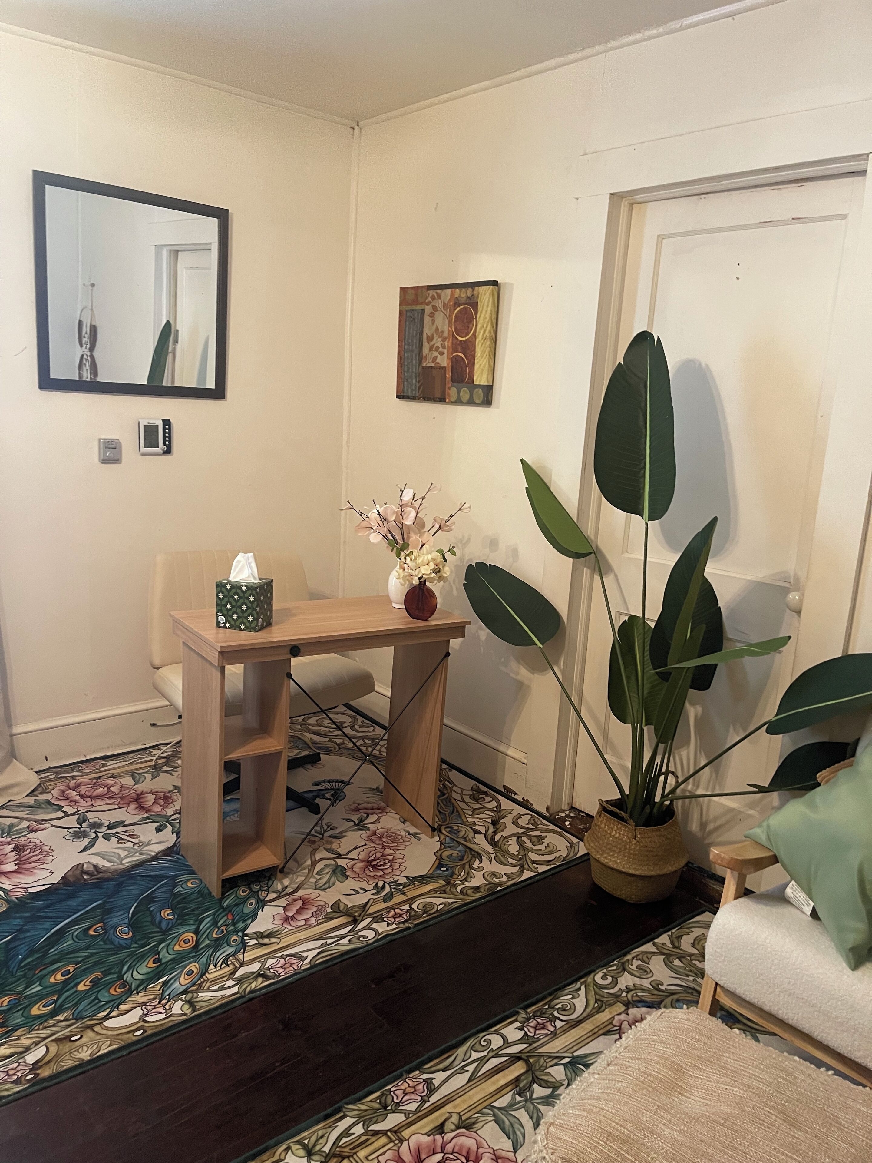 Una habitació amb un escriptori de fusta, una cadira, una planta gran en test, una taula petita amb un ram de flors, un sofà i una porta.