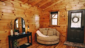 A cozy room with wooden walls, a round couch, a black console table with a mirror, and a door with a wreath.