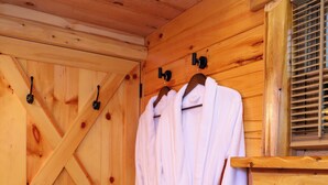 A wooden cabin interior with a wooden door, a window with blinds, and two white bathrobes hanging on hooks.