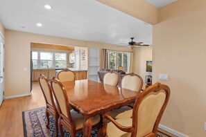 A dining room with a large wooden table and six chairs, a ceiling fan, and a view of the kitchen and garden through large windows. - Appleview - Walk to Village/Lake, Lake View, Hot Tub, BBQ (Big Bear Lake)