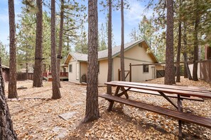 A cabin surrounded by tall pine trees, a picnic table, and a covered area with a roof. - Leaf & Needle | Single-Story Cabin, Fenced Yard, Wood Stove & Near The Village (Big Bear Lake)