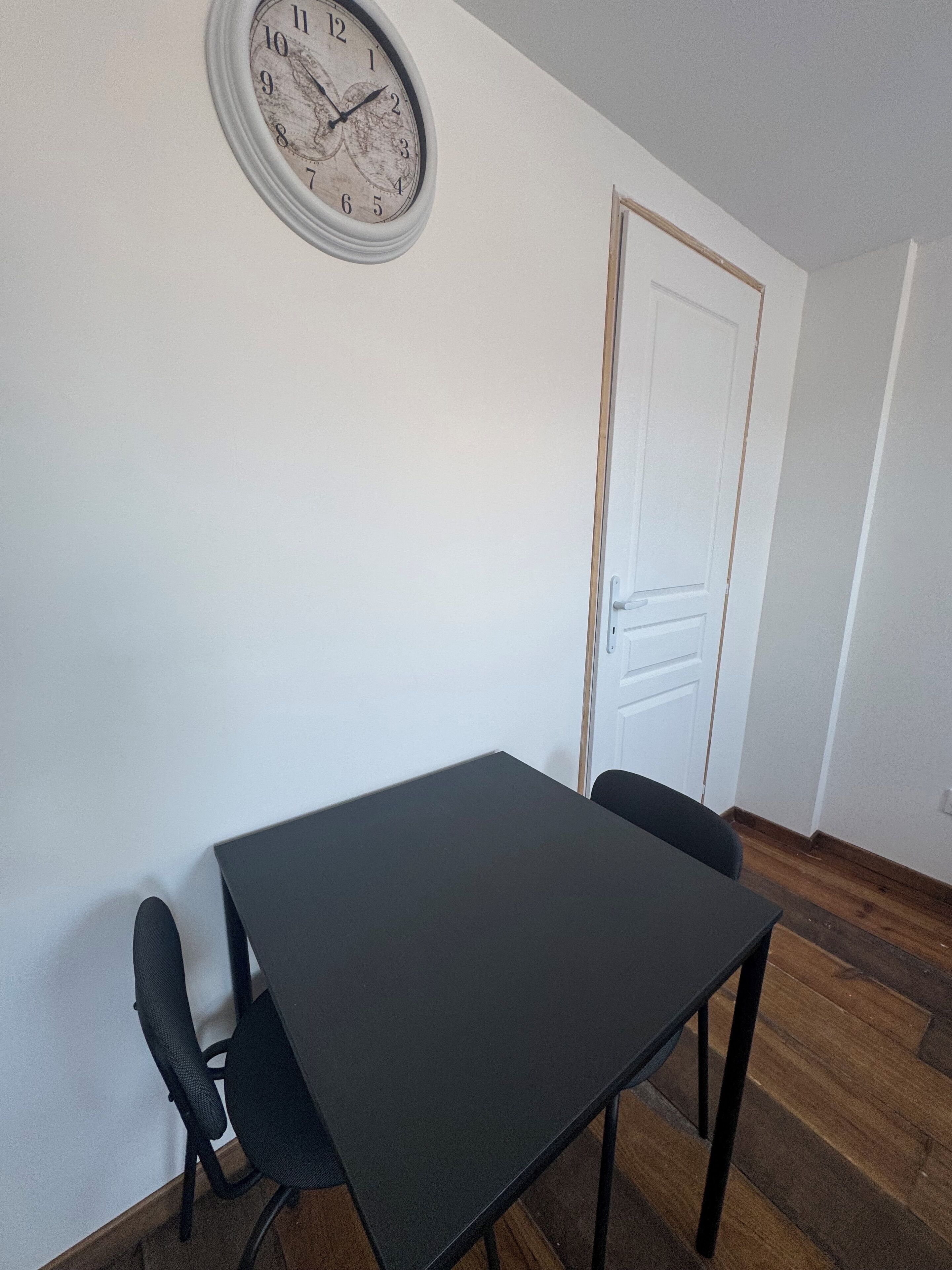 Ett rum med ett mörkt trägolv, en vit dörr, en rund väggklocka och ett svart bord med två stolar.