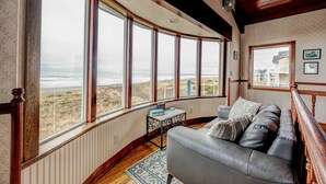 A room with a large bay window overlooking a beach, a grey sofa, a small table with books, and a patterned rug. - Serenity at South Beach (Westport)