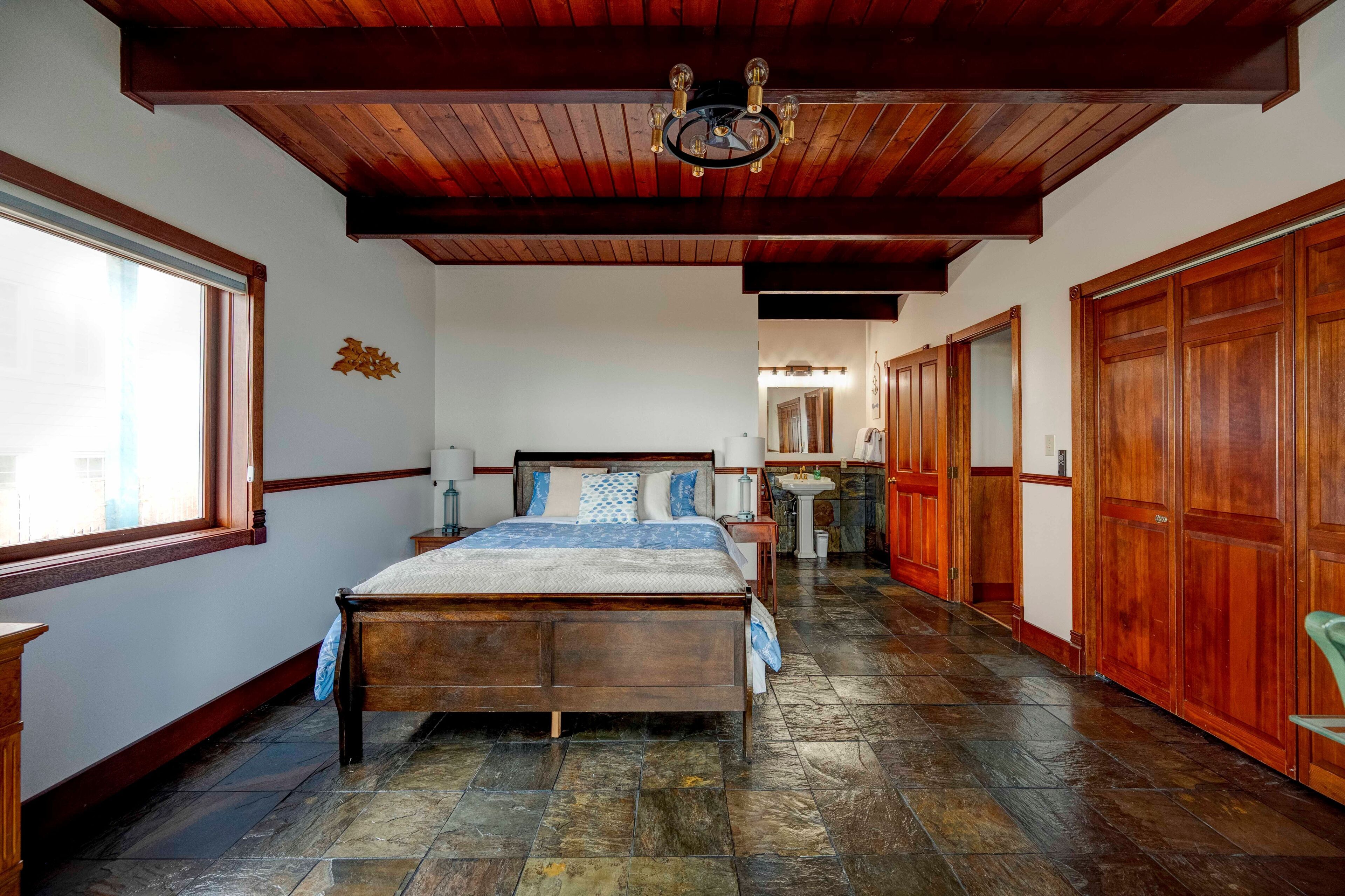 A bedroom with a wooden bed, stone flooring, a wooden ceiling, and a window.