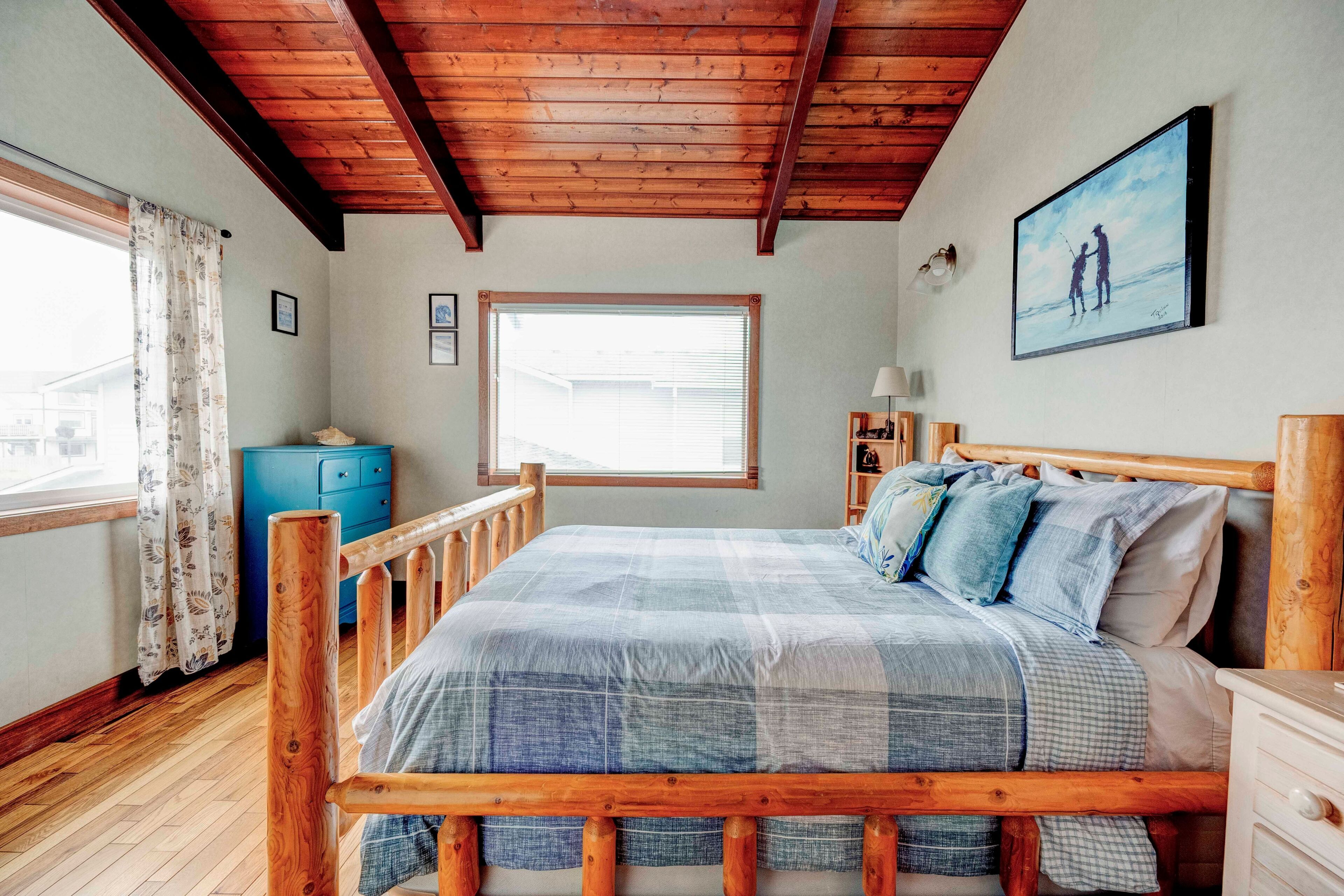 Un dormitorio con estructura de cama de madera, un ventanal grande, un aparador azul y un cuadro en la pared.