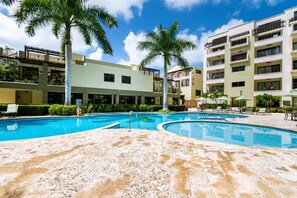 Une piscine dotée d’une terrasse ensoleillée, entourée de palmiers et bordée d’un immeuble à plusieurs étages avec des balcons.