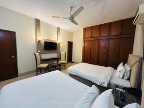 Ein Hotelzimmer mit zwei Einzelbetten, einem Schreibtisch mit Stuhl, einem an der Wand montierten Fernseher und einem Kleiderschrank.