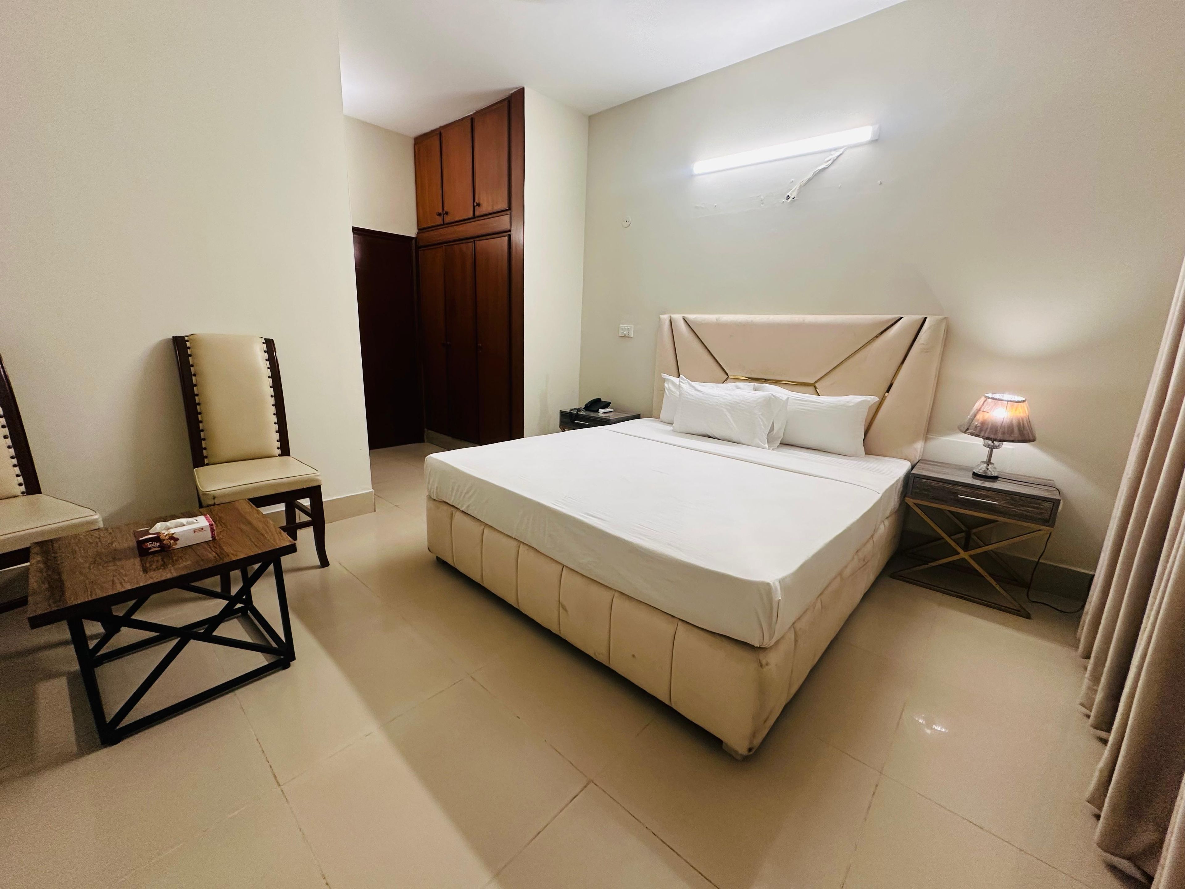 A bedroom with a large bed, a chair, a small table, a nightstand with a lamp, and a wardrobe.