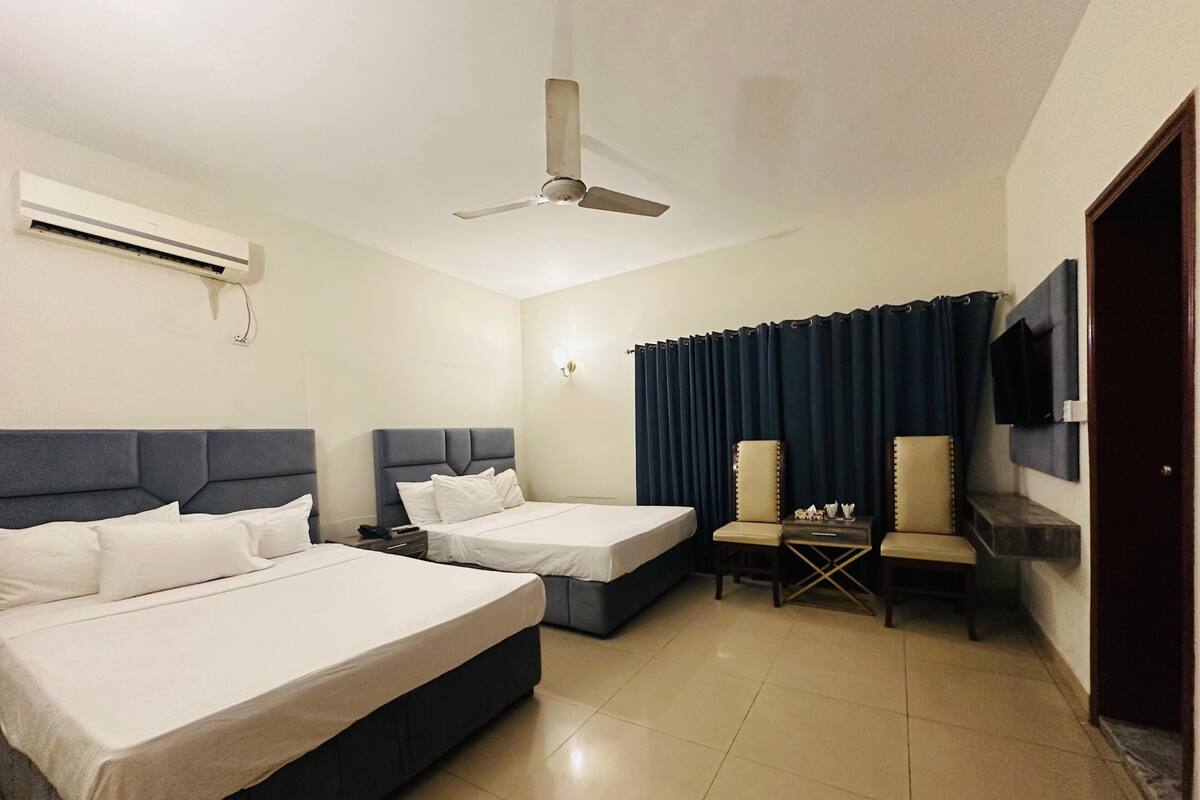 A hotel room with two beds, a ceiling fan, an air conditioning unit, a TV mounted on the wall, and a dark curtain.